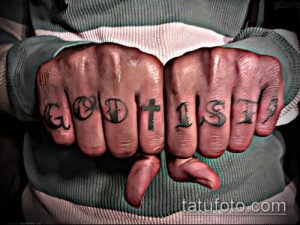 Фотография рук с татуировками на пальцах, на которых написаны слова "GOD IS" и изображен крест. Татуировки выполнены черным цветом.