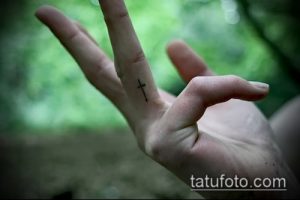 Фото тату: маленький черный крест на безымянном пальце руки. Простой и лаконичный рисунок татуировки.