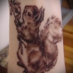 Фотография реалистичной татуировки белки, сидящей на ветке дерева. Детализированный рисунок татуировки с использованием коричневых и серых оттенков.