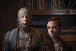 Фотография мужчины и женщины с татуировками, покрывающими их лица и тела. Они стоят рядом на фоне книжных полок.