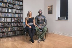 Фотография пары, где мужчина полностью покрыт татуировками в стиле биомеханики, а женщина с татуировкой на плече. Они сидят в комнате с книжными полками.