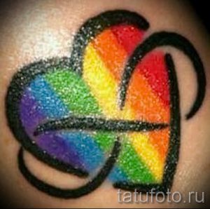 Цветная татуировка на руке с изображением абстрактного символа, заполненного радужными цветами и обведенного черным контуром. Эффектный и необычный рисунок татуировки.
