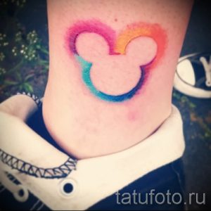 Фото акварельной тату на лодыжке с силуэтом головы Микки Мауса, выполненным в радужных цветах.