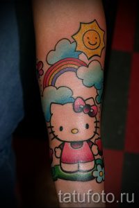 Большая татуировка на руке с изображением Hello Kitty, радуги, солнца и облаков. Яркий и милый рисунок татуировки.