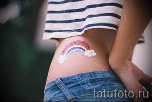 Фото тату радуги с облаками на женском боку. Небольшой рисунок татуировки выполнен в ярких пастельных тонах, создавая нежный и позитивный образ.