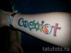 Фото тату надписи 'Coexist' с символами различных религий (ислам, иудаизм, христианство, буддизм, индуизм). Расположена на предплечье.