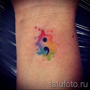 Фото тату в виде акварельной точки с запятой, выполненной в радужных цветах на запястье. Символ надежды и продолжения жизни.