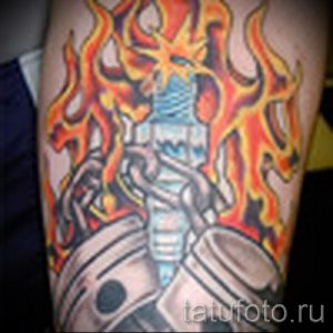 Фото тату с изображением пылающего двигателя и руки, держащей свечу зажигания. Рисунок выполнен в ярких оранжевых и синих тонах.