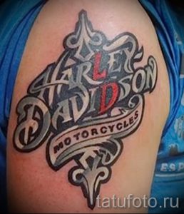 Фото тату логотипа Harley-Davidson на плече. Черно-серый рисунок, стилизованный шрифт, лента с надписью, классический дизайн.