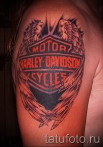 Фото тату логотипа Harley-Davidson на плече с добавлением крыльев и огненного фона. Яркий и динамичный рисунок.