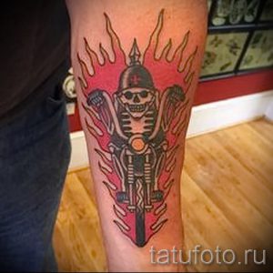 Фото тату: мотоцикл с черепом в шлеме, объятый пламенем, на руке. Использованы цвета: черный, красный и оранжевый. Детализированный рисунок татуировки.