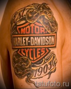 Фото тату: логотип Harley-Davidson с изображением мотоциклетных деталей на плече. Использованы цвета: черный, оранжевый, серый и белый. Детализированный рисунок татуировки.