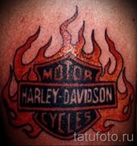 Фото тату: логотип Harley Davidson, окруженный пламенем, яркие красные и оранжевые цвета, динамичный рисунок, символика страсти и свободы.