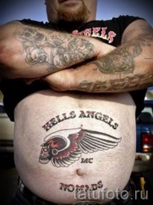 Фото татуировки логотипа Hells Angels на животе байкера. Детализированный рисунок с использованием черных и красных тонов. Символ свободы и принадлежности к мотоциклетному клубу.
