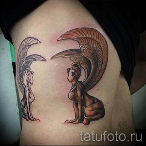 Фото тату на боку с изображением двух женских фигур с крыльями, выполненное в приглушенных тонах с использованием черного, серого и красного цветов.