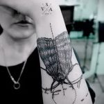 Фото тату мотылька на руке. Черно-белый рисунок в стиле лайнарт с тонкими линиями и акцентом на детали. Эскиз татуировки с добавлением текстовых элементов.