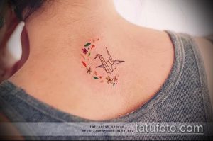 Небольшое фото тату на лопатке с изображением бумажного журавлика, окруженного яркими цветами в стиле минимализм.