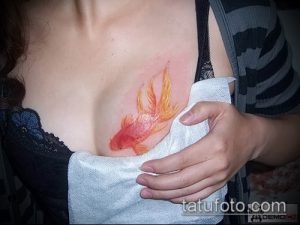 Фото тату золотой рыбки на груди. Рисунок выполнен в акварельном стиле, с использованием ярких оранжевых и желтых оттенков. Красивый и необычный эскиз тату.