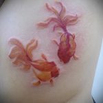 Фото акварельной татуировки с двумя золотыми рыбками на боку. Нежные пастельные тона, размытые контуры, создающие эффект акварельной живописи.