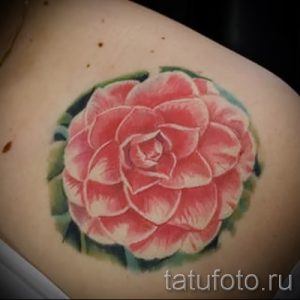 Фото тату крупной розовой камелии с зелеными листьями на плече. Яркий и сочный рисунок, выполненный в технике акварели с реалистичными деталями.