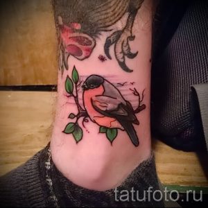 Фото тату птички, сидящей на ветке с листьями. Мягкие цвета, детальная проработка, нежный и романтичный дизайн на лодыжке.
