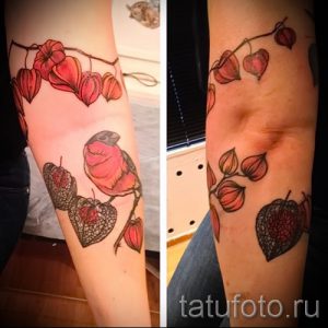 Фото тату на предплечье: красная птица сидит на ветке с фонариками. Детализированный рисунок татуировки в акварельном стиле с использованием красных и черных оттенков.