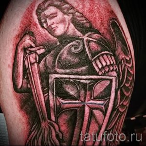 Фото тату на плече: изображение архангела Михаила с мечом, щитом и крыльями. Детализированный рисунок татуировки в черно-серых тонах с красными акцентами.