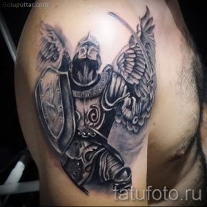 Фото тату на плече: изображение воина-ангела в доспехах с мечом, щитом и крыльями. Детализированный рисунок татуировки в черно-серых тонах.