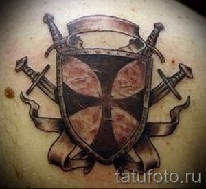 Фото тату: стилизованный щит с крестом, окруженный скрещенными мечами и лентами. Детализированный рисунок татуировки в черно-серых тонах.
