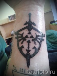Фото тату на запястье с изображением стилизованного щита и меча из игры The Legend of Zelda. Черно-белый рисунок татуировки в минималистичном стиле.