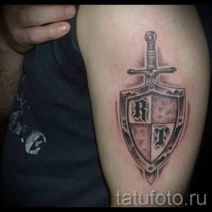 Фото тату на руке с изображением щита, украшенного буквами 'R' и 'T', пронзенного мечом. Рисунок выполнен в черно-серых тонах.