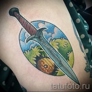Фото тату на руке: меч, пронзающий круглый пейзаж с небом, облаками и деревьями. Цветная татуировка с яркими красками и детальной проработкой.