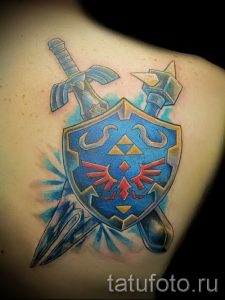 Фото татуировки на плече с изображением меча и щита из игры Zelda. Цветной рисунок татуировки с использованием ярких оттенков синего и красного.