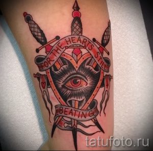 Фото татуировки в стиле олд скул на руке с изображением сердца, пронзенного кинжалами, и всевидящего ока. Яркий рисунок татуировки с четкими контурами.