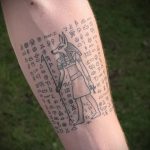 Фото тату Анубиса на руке, выполненное в минималистичном стиле с использованием тонких линий и иероглифов. Детализированный рисунок головы бога.