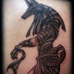 Фото тату Анубиса. Черно-белый рисунок в стиле blackwork изображает бога с головой шакала, держащего анкх и обвитого змеей.