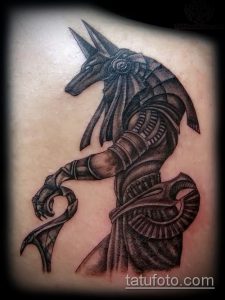 Фото тату Анубиса. Черно-белый рисунок в стиле blackwork изображает бога с головой шакала, держащего анкх и обвитого змеей.