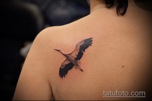 Фото небольшой татуировки аиста в полете на плече. Реалистичный рисунок с использованием серых и черных оттенков.