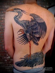 Фото тату аиста на спине мужчины. Реалистичный рисунок птицы с расправленными крыльями и волнами внизу.