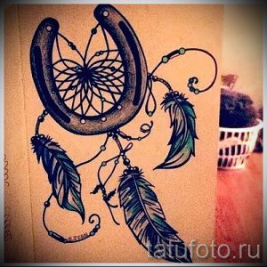 Фото тату подковы, украшенной ловцом снов и тремя перьями. Черно-серый рисунок татуировки с детальной проработкой элементов.