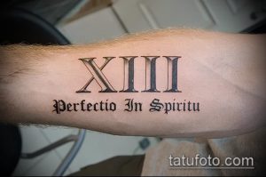 Фото татуировки на предплечье с римскими цифрами XIII и латинской фразой 'Perfectio In Spiritu'. Черно-серый дизайн, четкие линии, классический стиль.