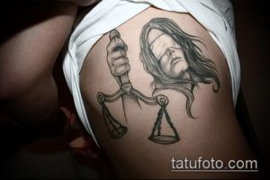 Фото тату Фемиды на бедре. Черно-серый рисунок татуировки с изображением богини правосудия с весами и повязкой на глазах.