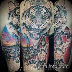 Фото тату на предплечье: свирепый тигр с мечом в пасти и гладиатор на фоне Колизея. Реалистичный рисунок татуировки в черно-серых тонах.