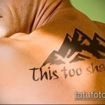 Фото тату с изображением гор и надписью "This too shall pass" на спине. Черно-белый рисунок татуировки, символизирующий надежду и веру.