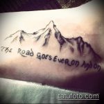 Фото тату на запястье с изображением гор и надписью "The road goes ever on and on". Черно-белый рисунок татуировки, символизирующий путешествие и бесконечность.