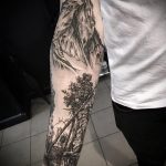 Фото реалистичной татуировки с изображением горного пейзажа и леса на руке. Детализированная проработка и черно-белая гамма.