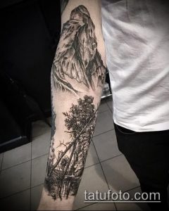 Фото реалистичной татуировки с изображением горного пейзажа и леса на руке. Детализированная проработка и черно-белая гамма.