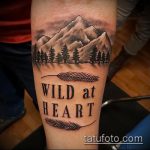 Фото тату горного пейзажа с надписью "Wild at Heart" на предплечье, выполненного в черно-серых тонах с использованием реалистичной прорисовки.