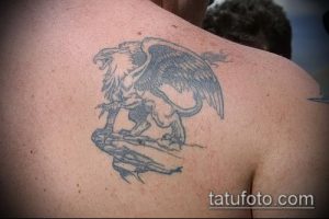 Большая черно-серая татуировка на спине, изображающая грифон, нападающего на воина. Детализированный рисунок с проработкой перьев и доспехов.
