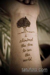Фото тату на предплечье: небольшое дерево и вдохновляющая цитата "i climbed the tree to see the world". Эскиз татуировки выполнен в коричневых тонах.